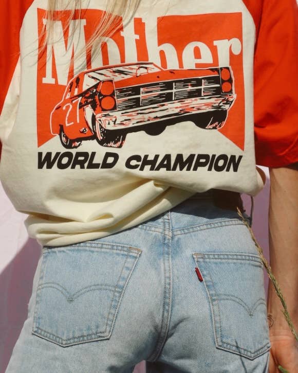 Person wearing a shirt with a graphic of a car and 'Mother World Champion' text, standing against a pink background.
