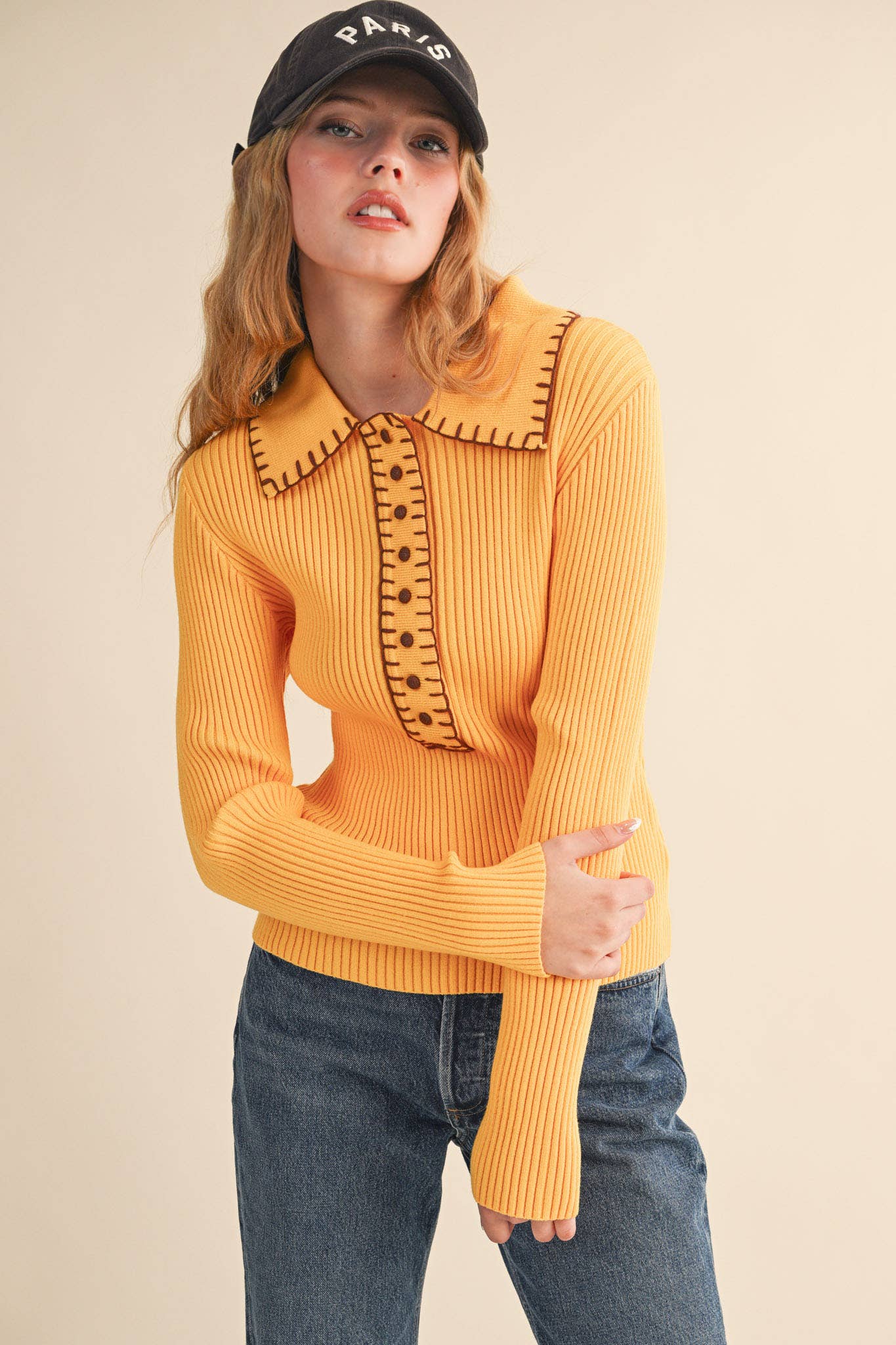 Woman wearing a mustard yellow sweater with black collar details and a black cap on a beige background
