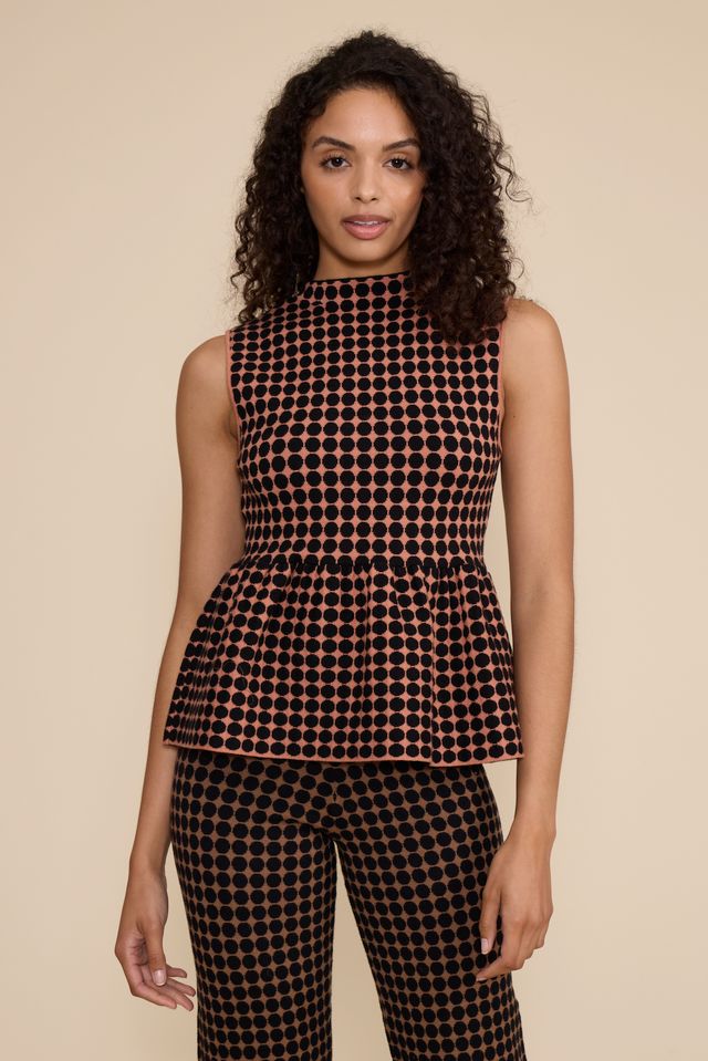 A woman modeling a sleeveless peplum top and matching pants set in a black and copper-orange checkered pattern, set against a solid beige studio background.
