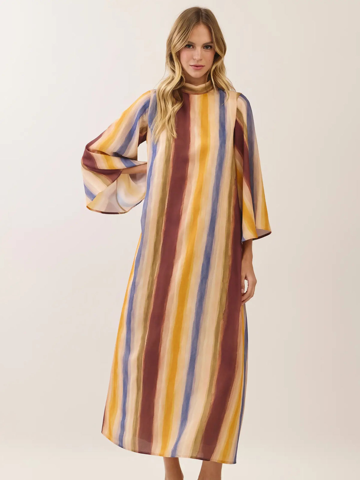 A woman modeling a long-sleeved midi dress with vertical stripes in mustard yellow, brown, blue, and cream against a plain light beige studio background.