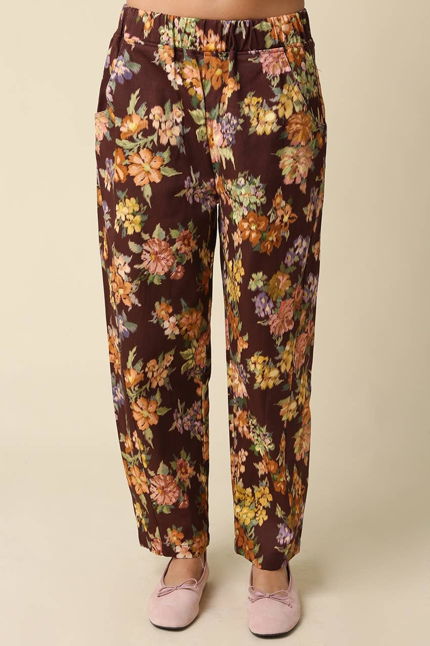 A person wearing dark brown floral pants with a multicolor print of orange, yellow, and purple flowers, paired with light pink slip-on shoes, against a plain beige background.