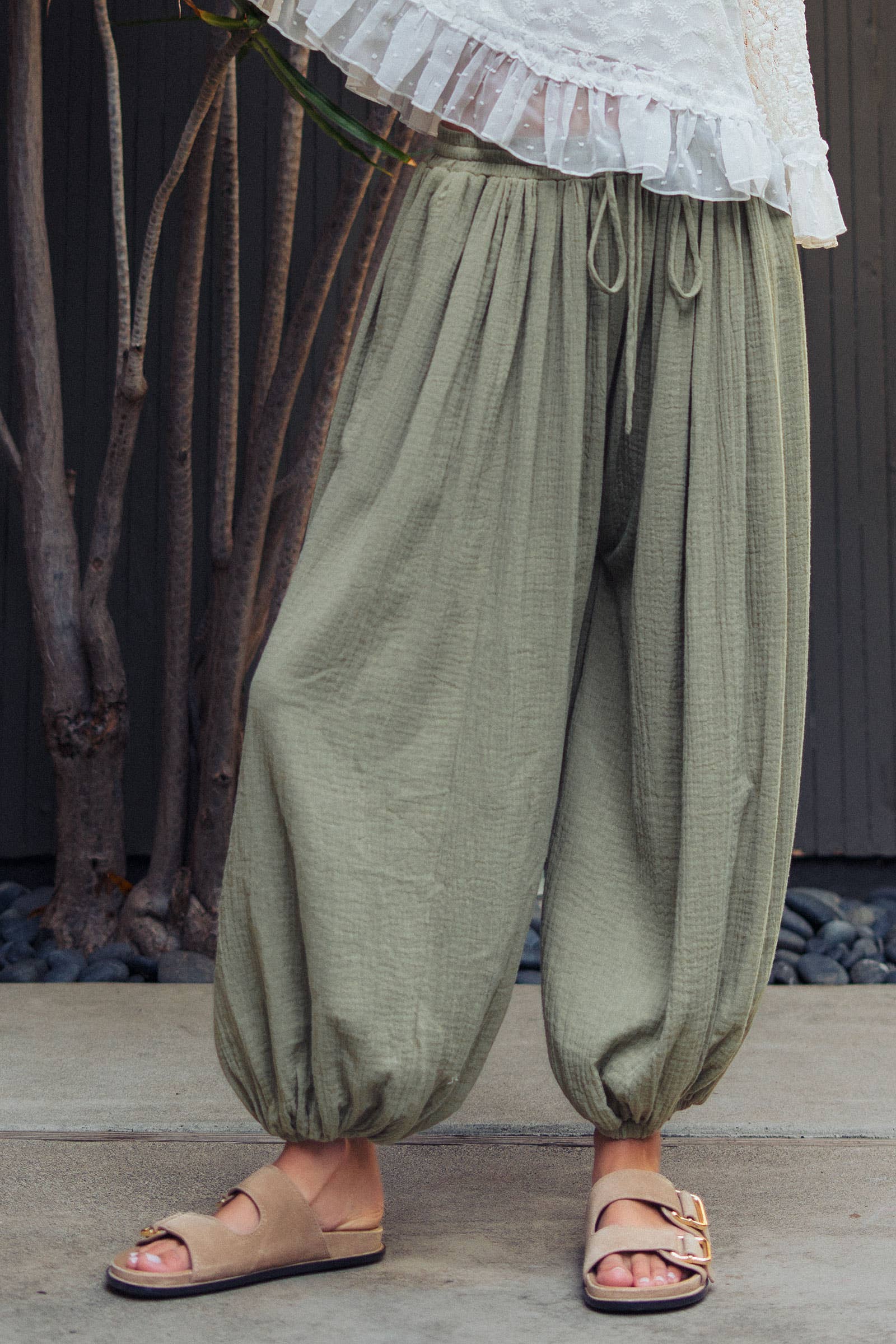 Person wearing olive green harem pants with a white top and sandals, standing against a natural background.