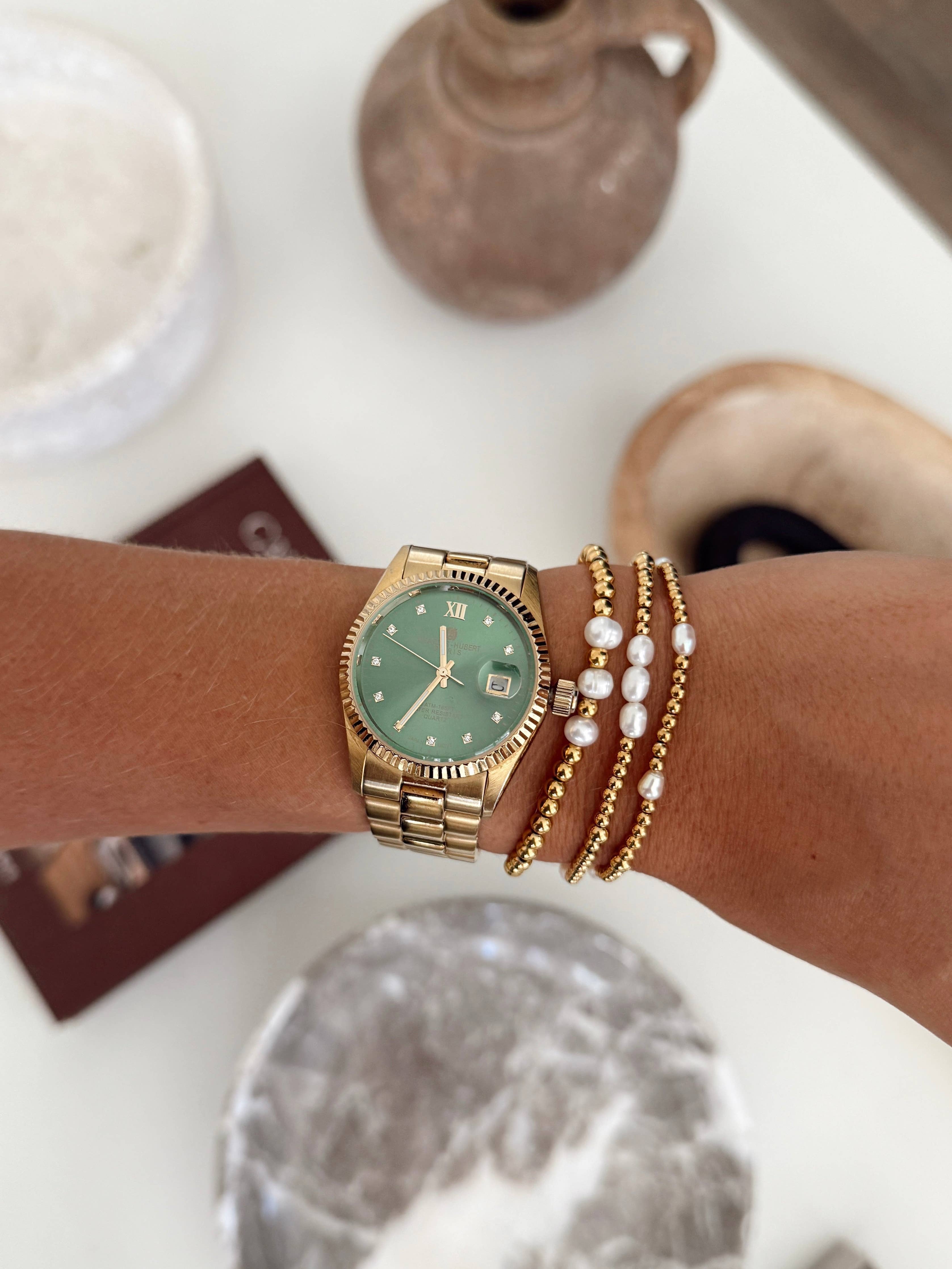 Gold watch with a green face on a wrist, surrounded by pearl bracelets, with a blurred background.
