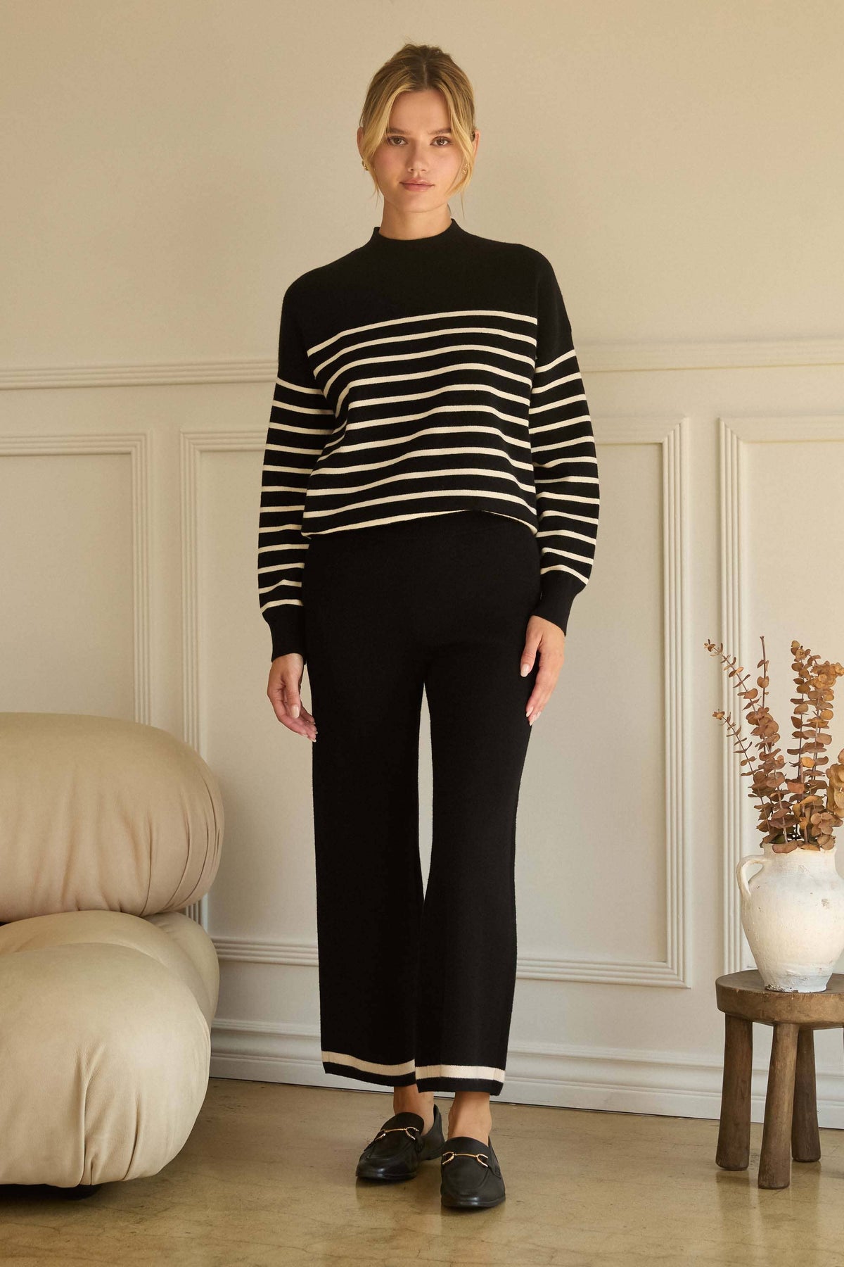 A woman stands indoors wearing a black knit mock-neck sweater with horizontal cream stripes on the sleeves and upper chest, paired with black wide-leg cropped pants and black loafers. The room has cream paneled walls, a beige sofa on the left, and a small wooden side table with a vase on the right.