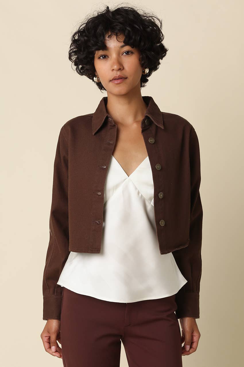 A model wearing a dark brown cropped button-up shirt jacket over a white blouse, paired with dark brown trousers, against a solid beige background.