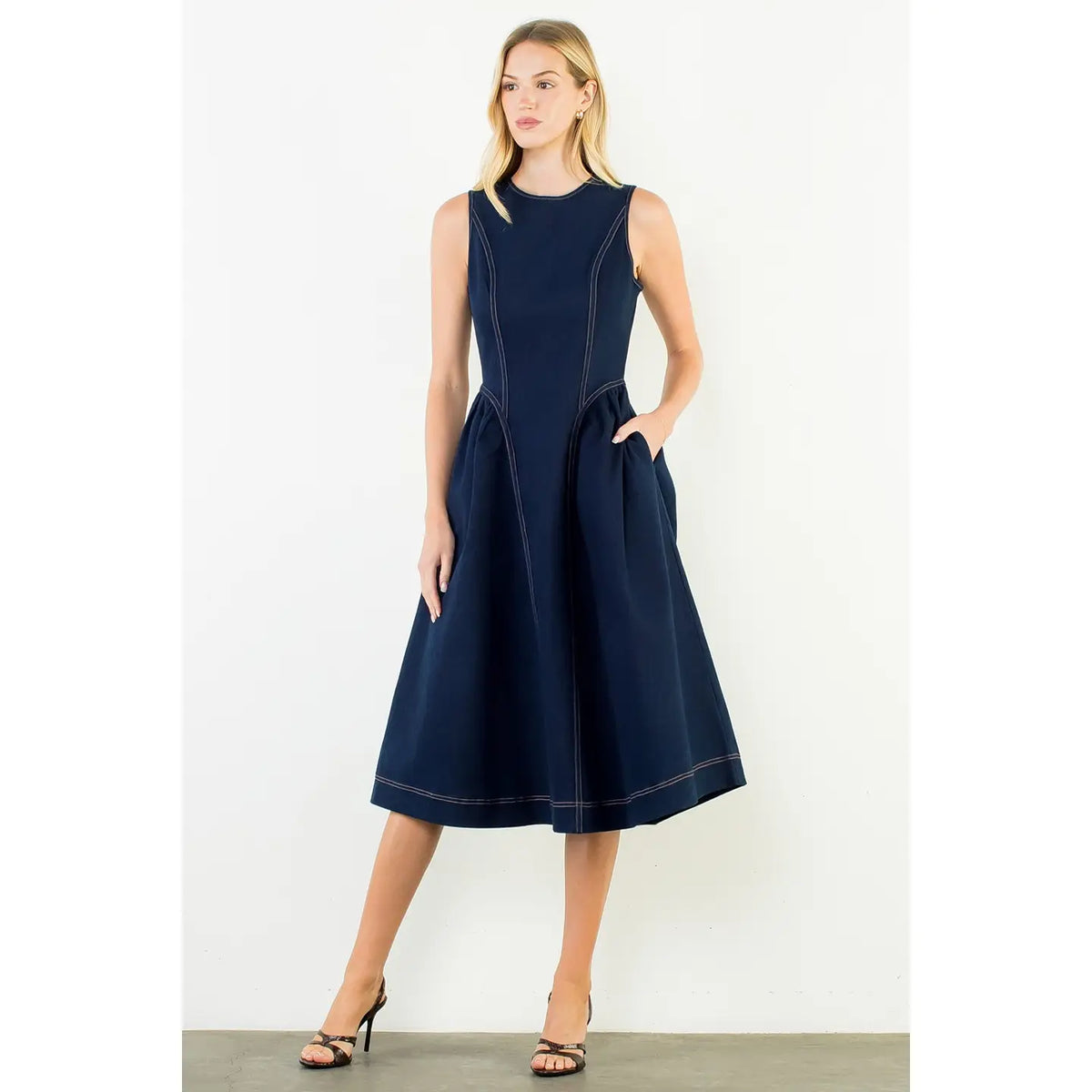 A woman modeling a sleeveless dark navy midi dress with contrast stitching, side pockets, and a flared skirt, standing against a plain off-white studio background.