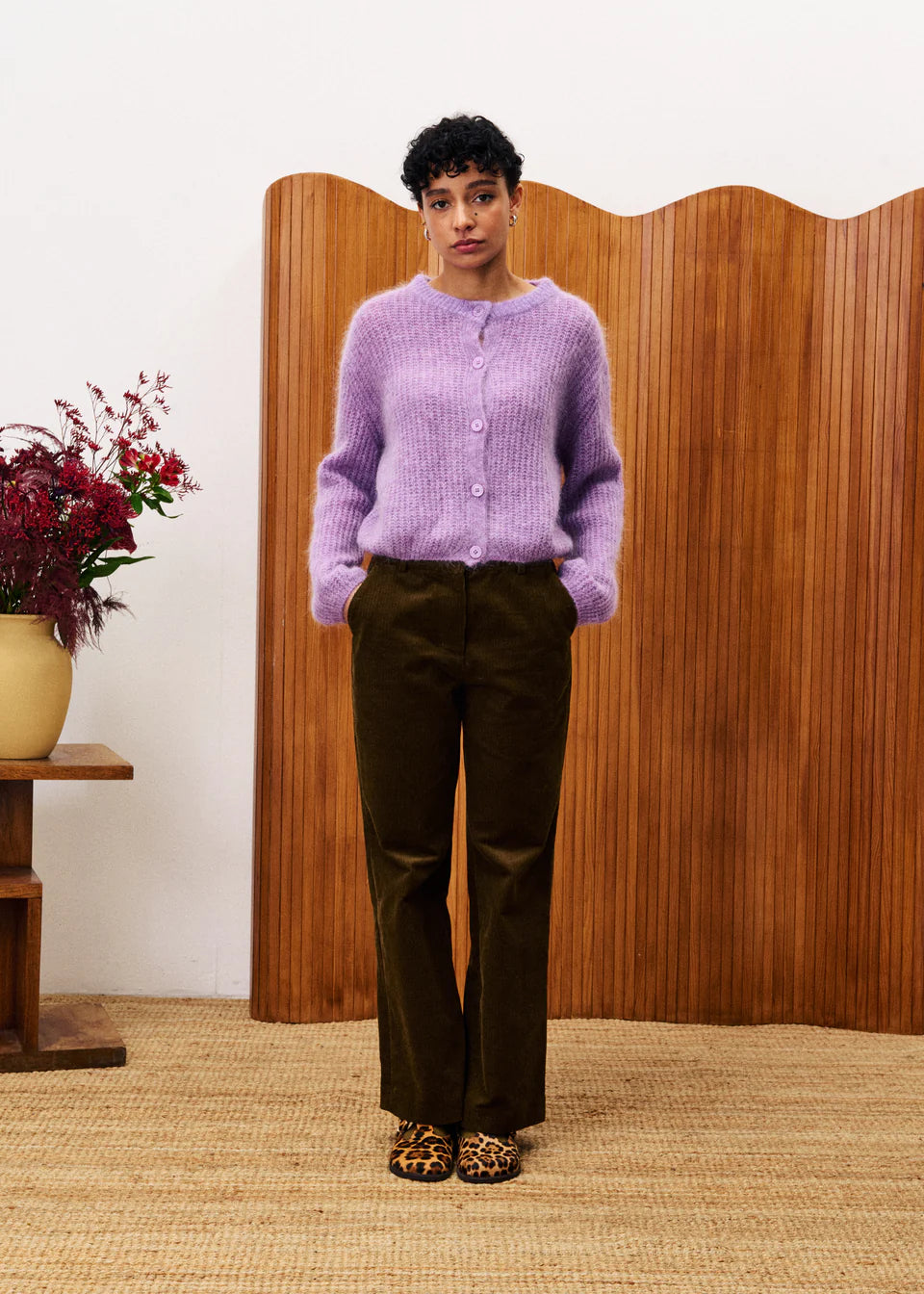 Person wearing a purple cardigan and dark pants standing in front of a wooden screen.