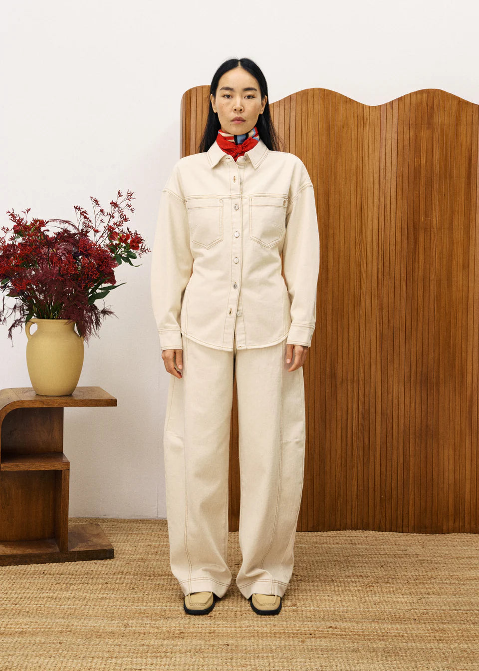 Person wearing a cream-colored outfit standing in front of a wooden screen with a vase of flowers on a table.
