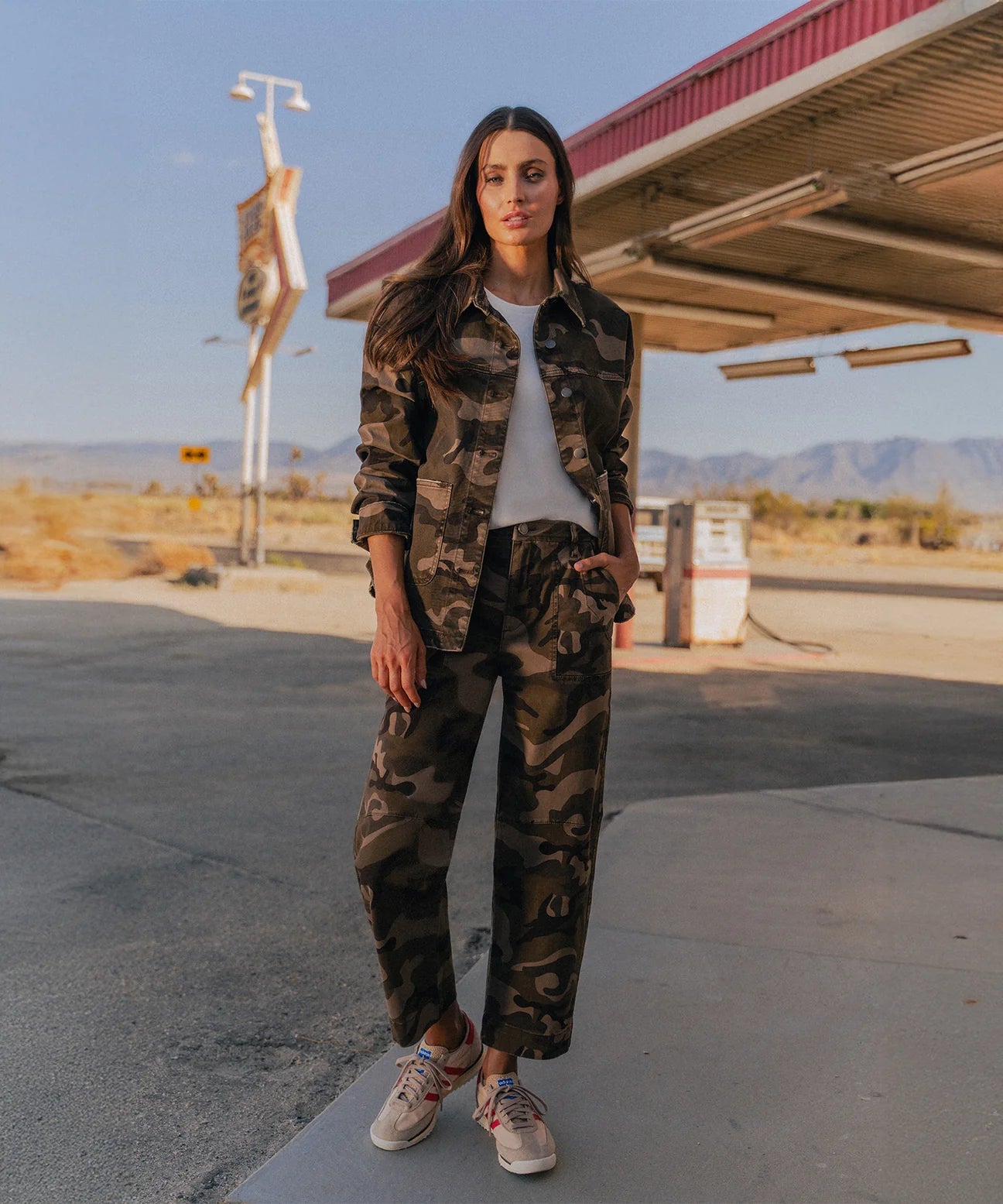 Person wearing a camouflage outfit standing in a desert-like setting with a gas station in the background.