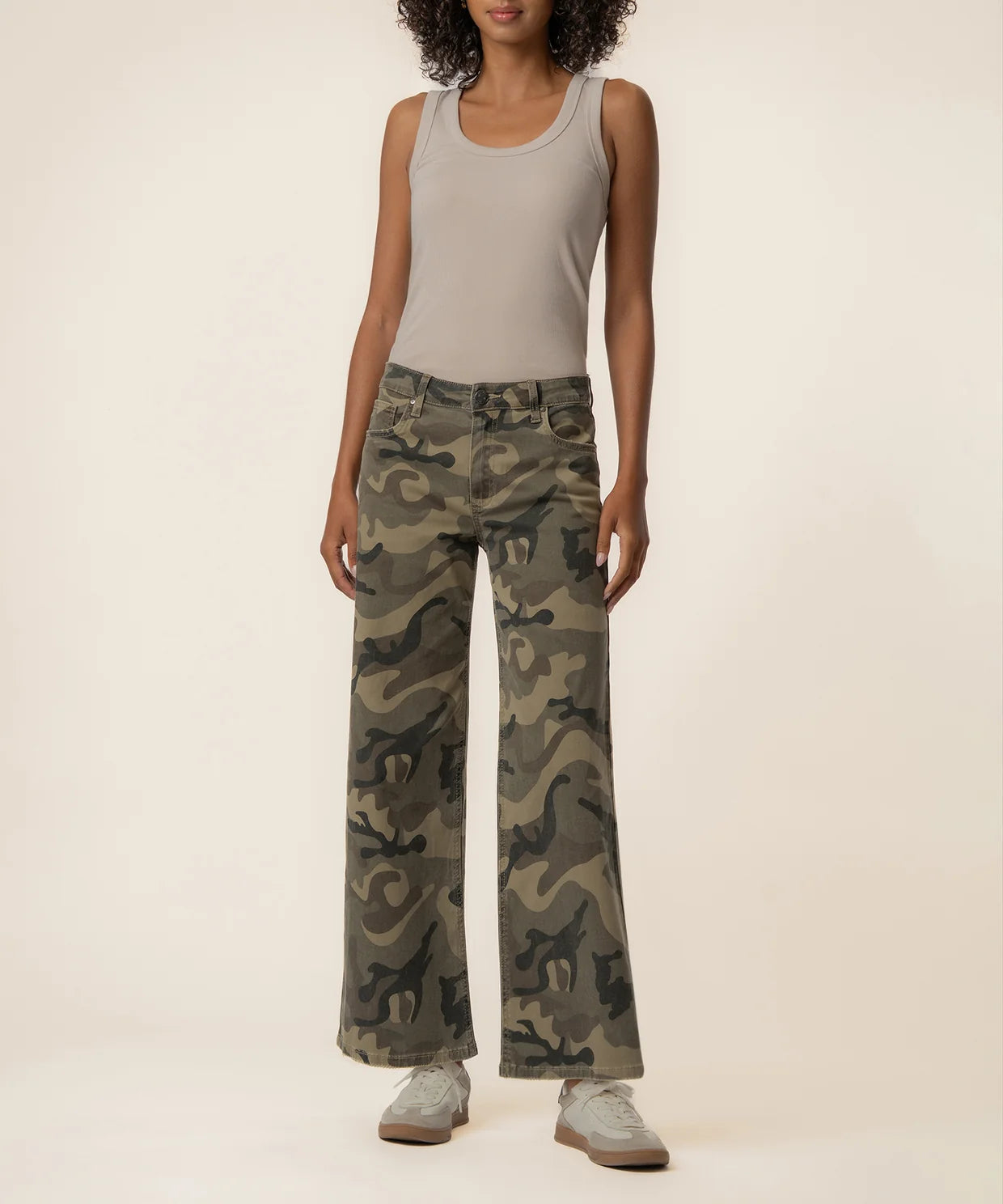 A model wearing a pair of wide-leg camouflage pants, styled with a light gray tank top and gray sneakers, standing against a solid beige studio background.