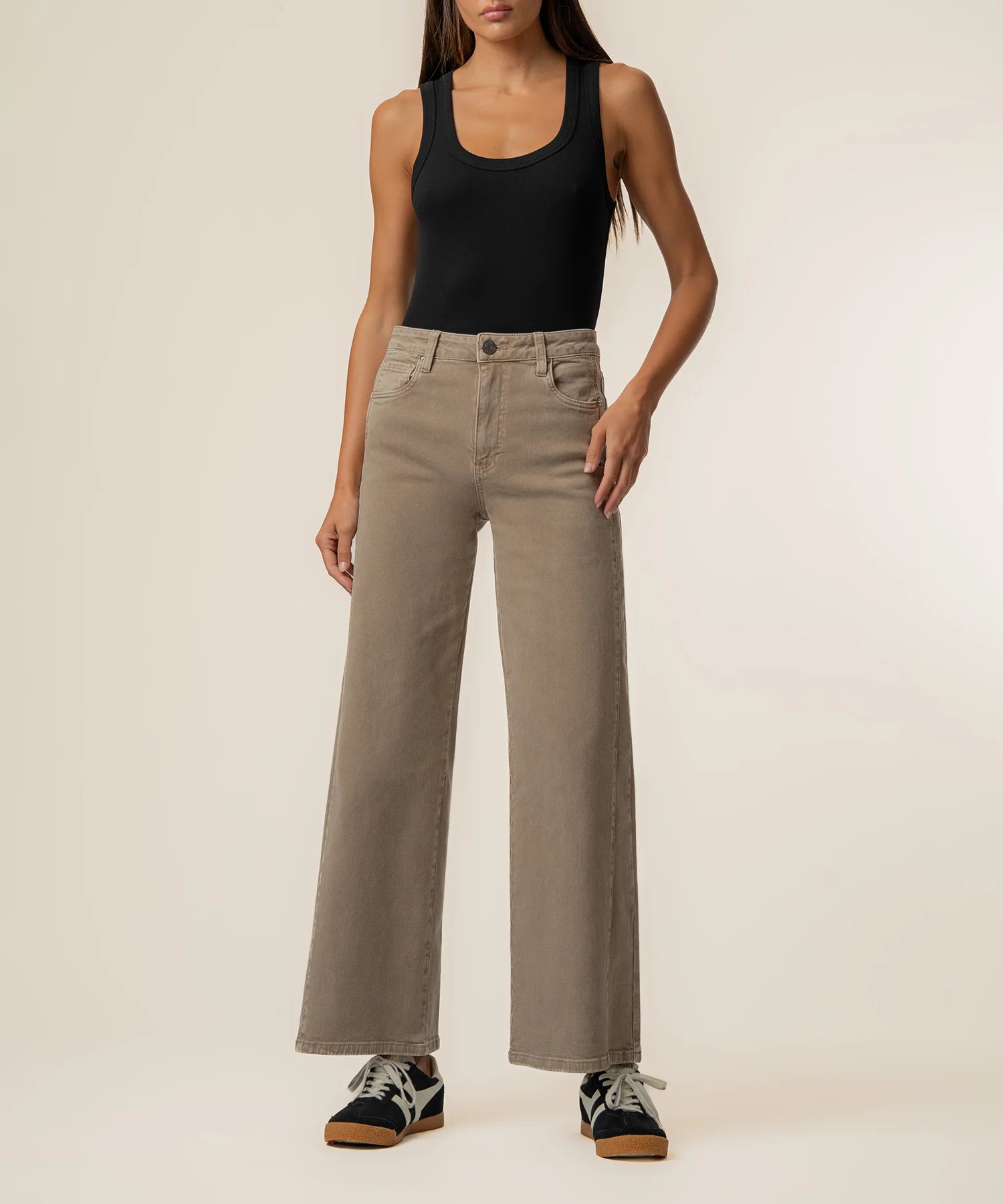 A model wearing a pair of high-waisted, wide-leg taupe jeans, paired with a black tank top and black sneakers, standing against a plain off-white studio background.