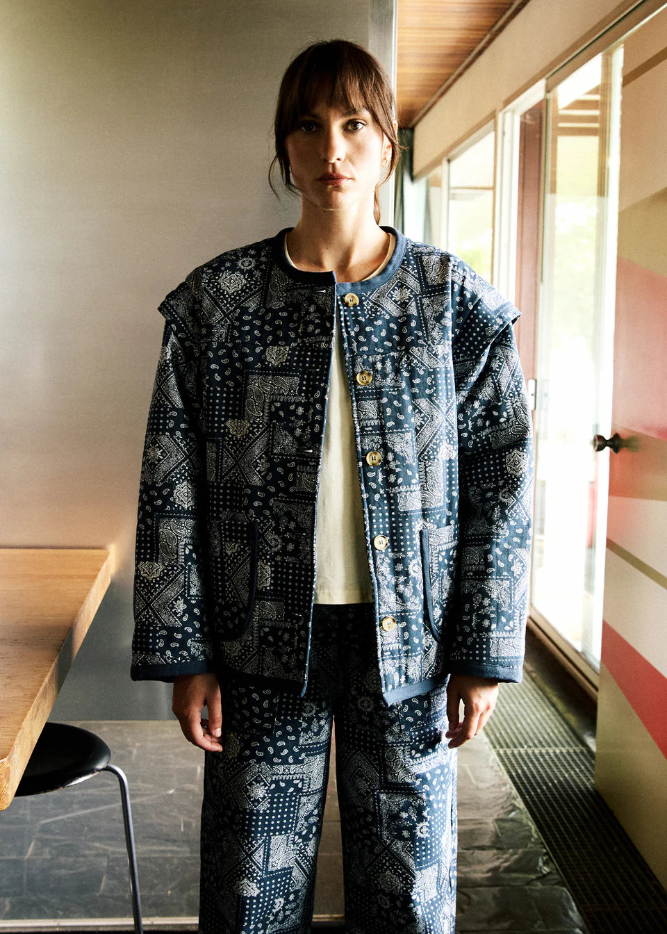 Person wearing a patterned jacket and pants standing indoors near a window.