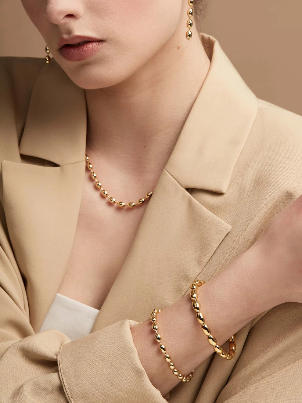 Close-up of a person wearing gold jewelry including a necklace, bracelet, and earrings against a beige background.