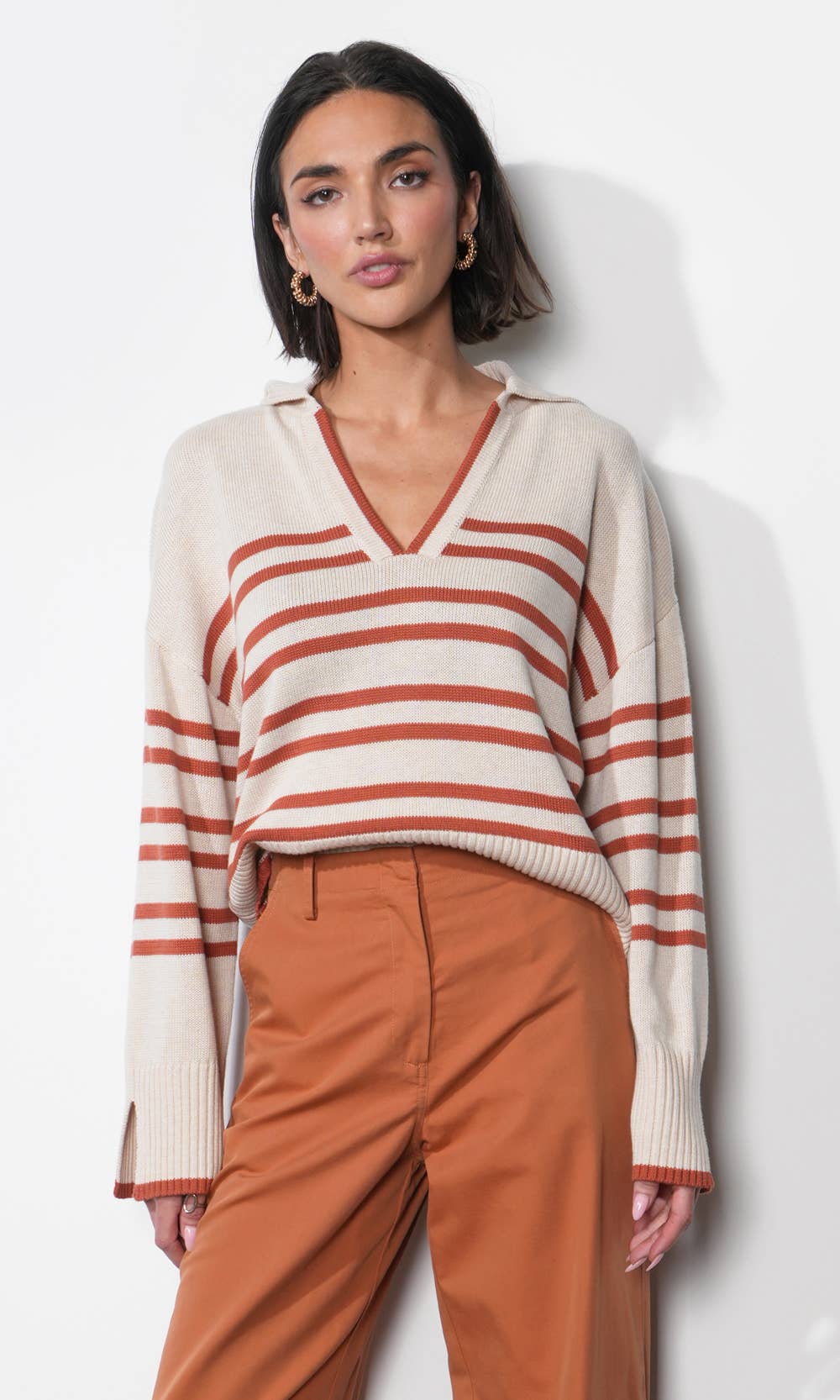 A woman modeling a cream knit sweater with rust-orange horizontal stripes and a V-neck, paired with rust-orange pants, against a light gray studio background.