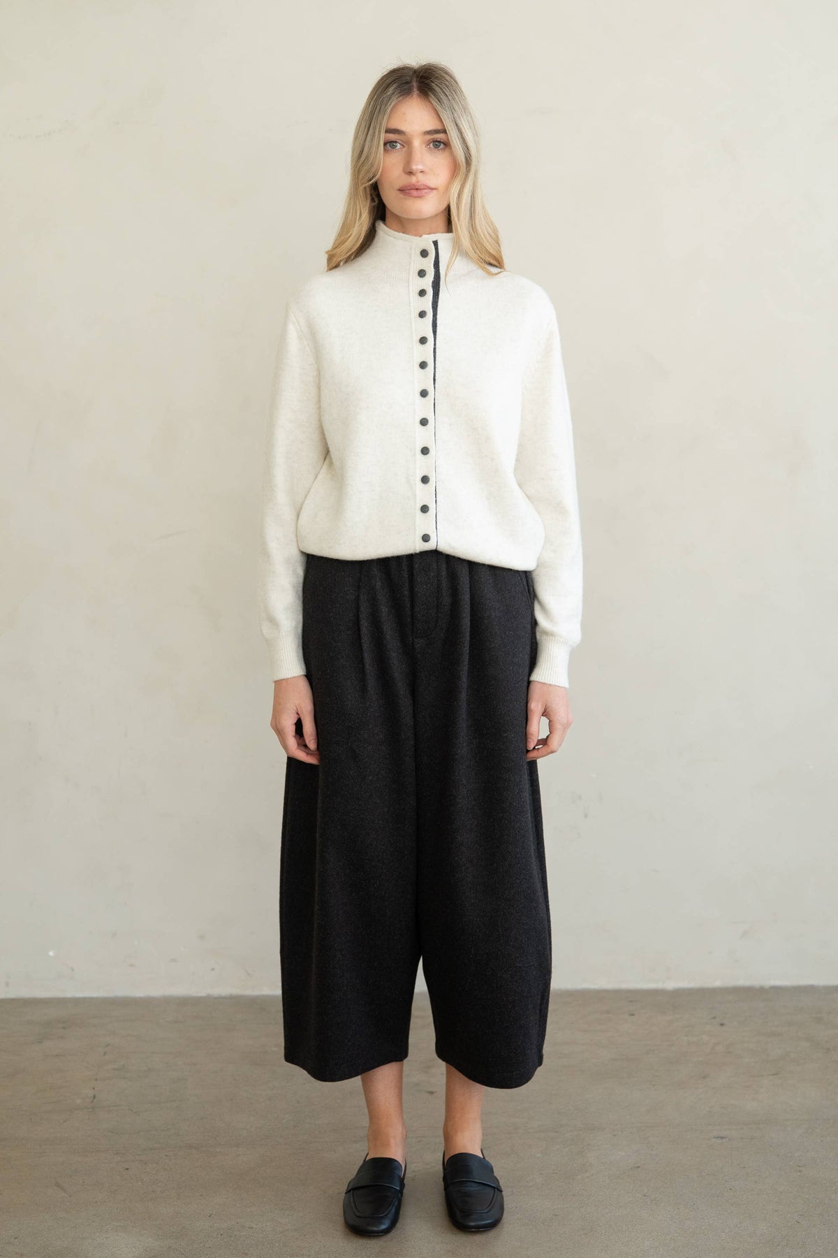 A woman stands against a plain light beige wall wearing a cream knit mock-neck cardigan with a central button placket, black wide-leg cropped pants, and black slip-on shoes.