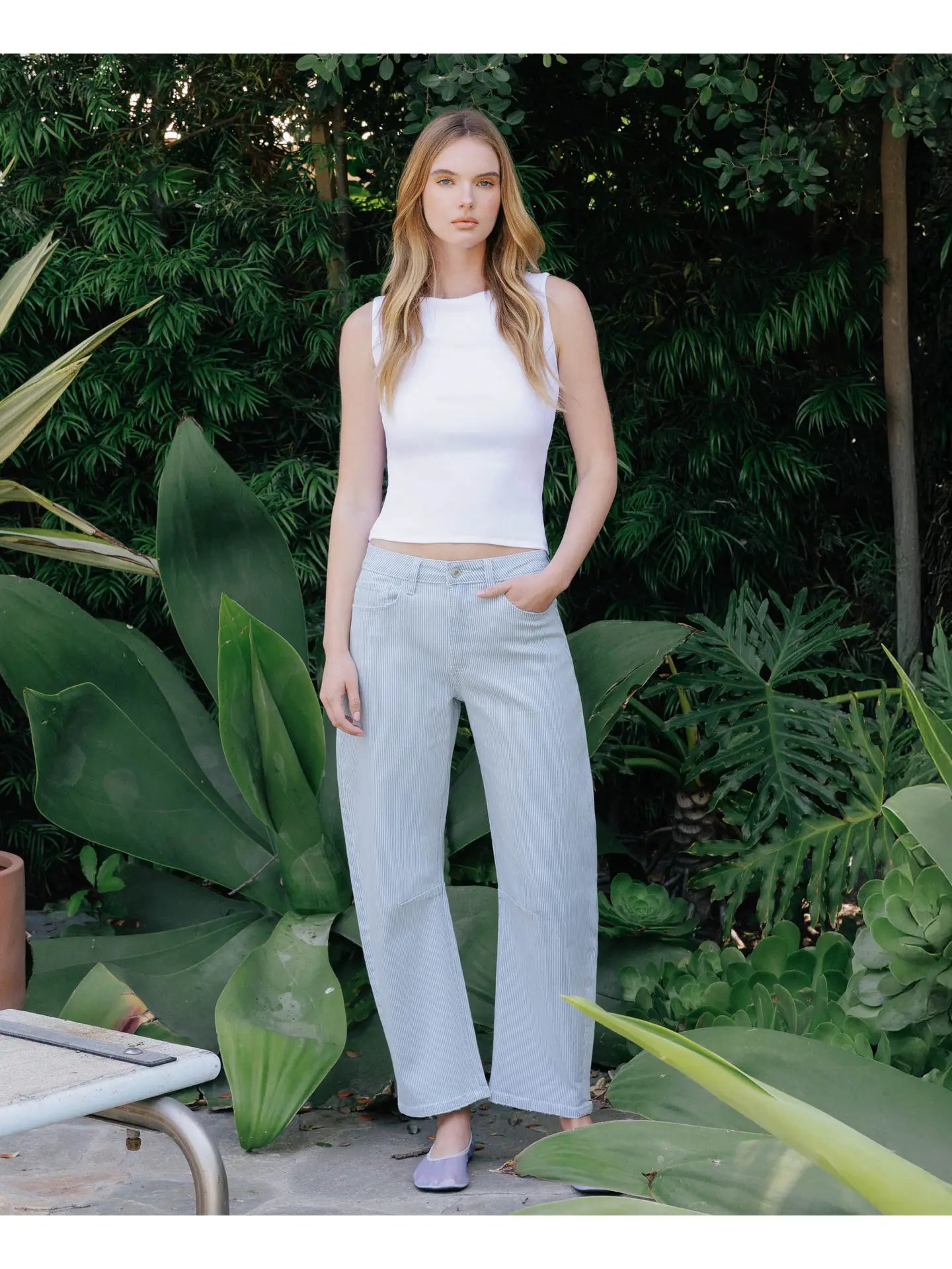 Woman wearing a white sleeveless top and light blue pants standing in a garden.