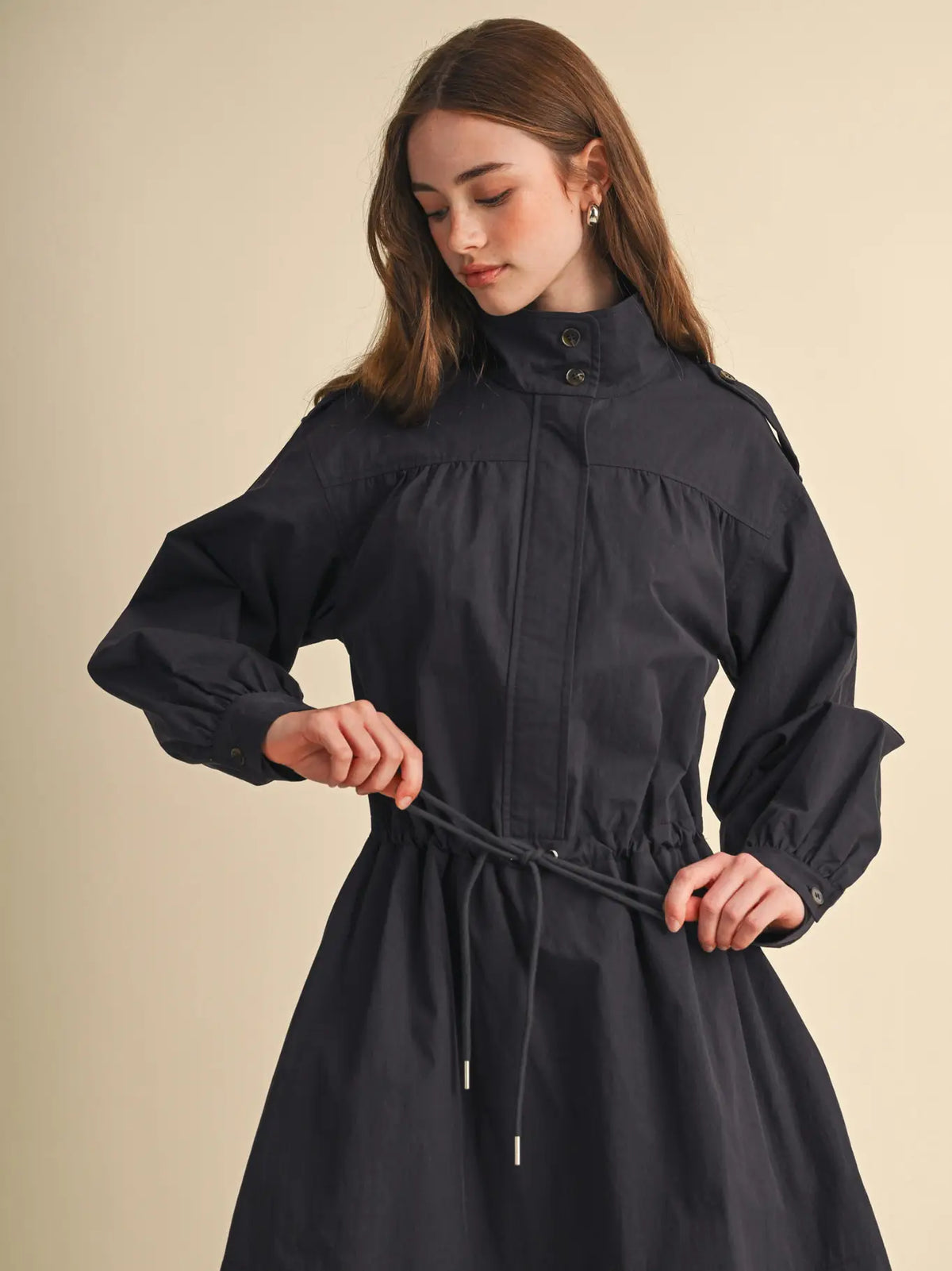 A woman wearing a dark charcoal gray high-neck long-sleeve dress with a drawstring waist, standing against a solid beige background.
