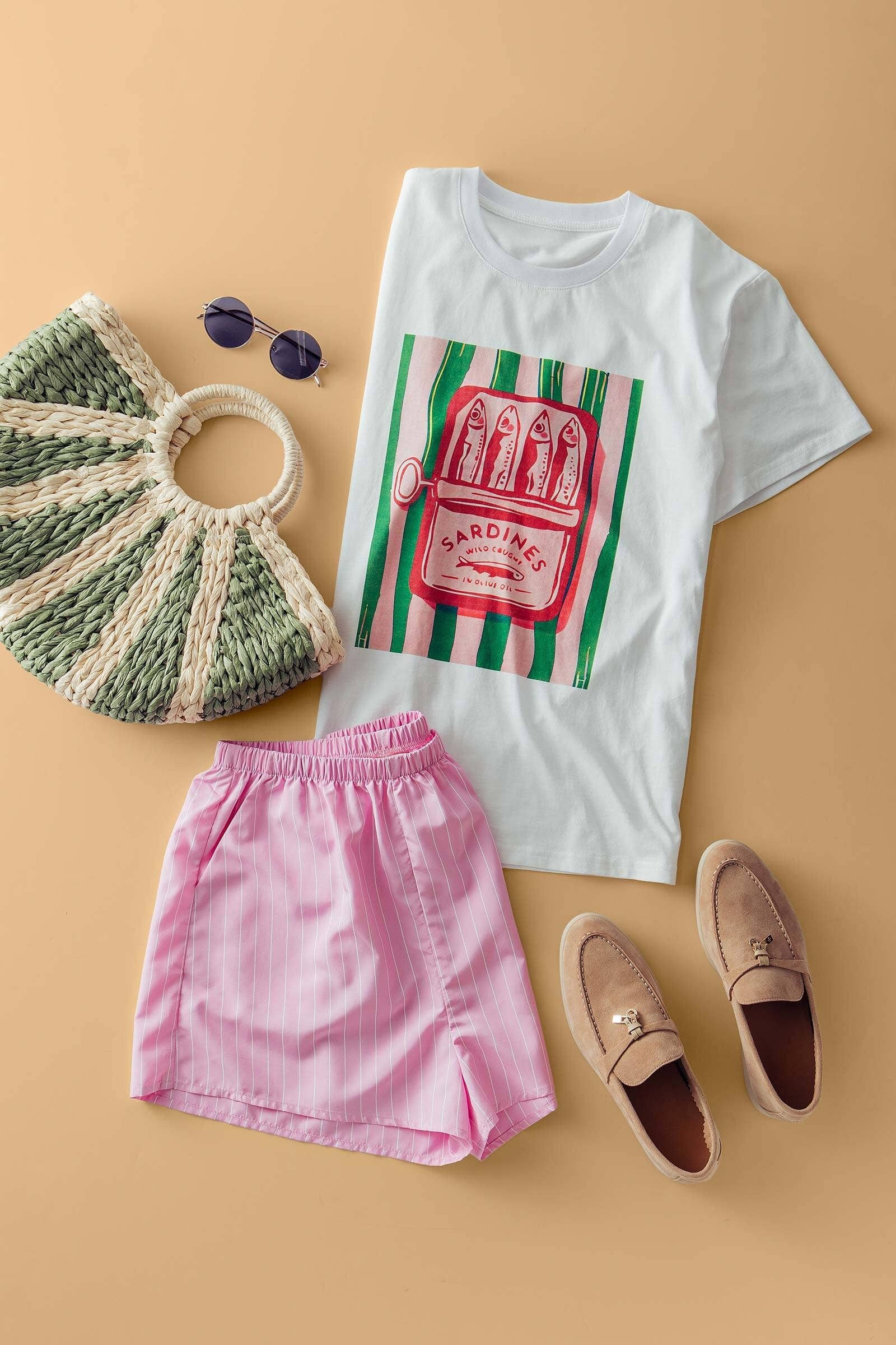 White t-shirt with graphic design, pink shorts, beige loafers, and a woven bag on a beige background