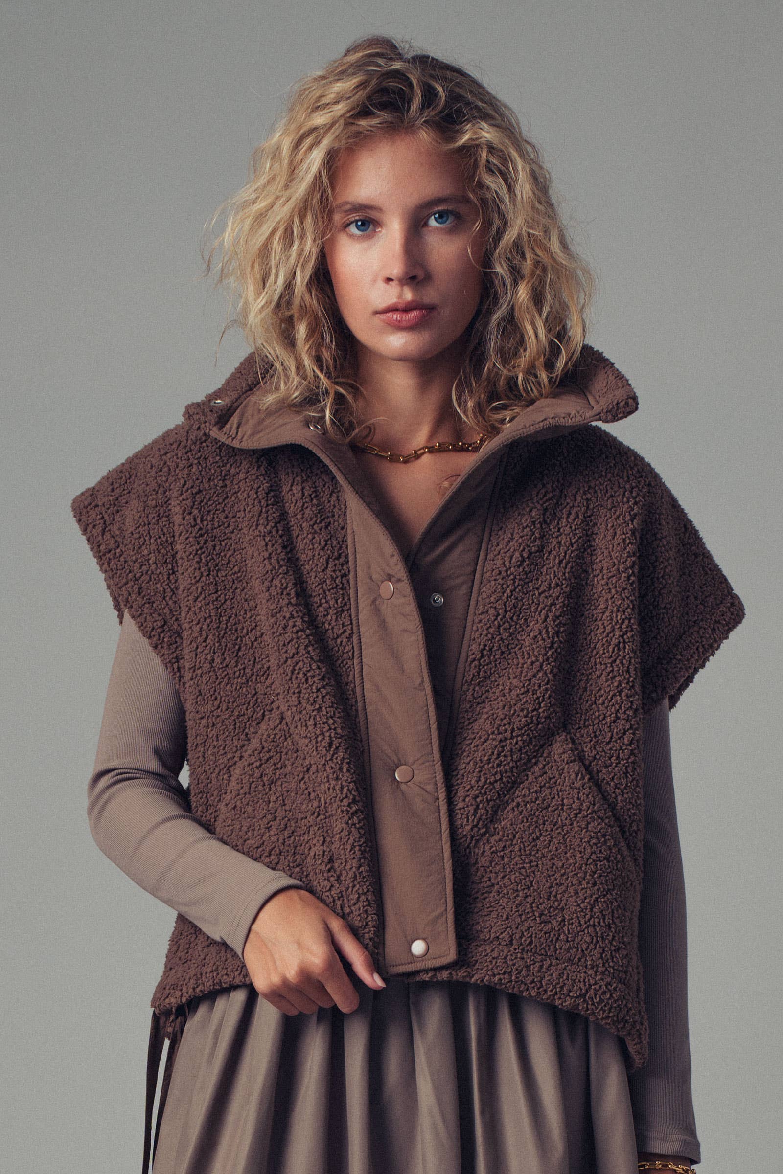 Woman wearing a brown knitted vest over a long-sleeve shirt against a gray background