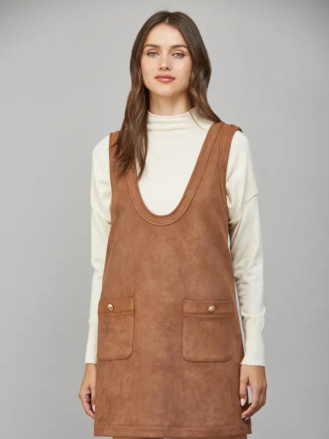 Woman wearing a brown suede pinafore dress over a white turtleneck on a gray background