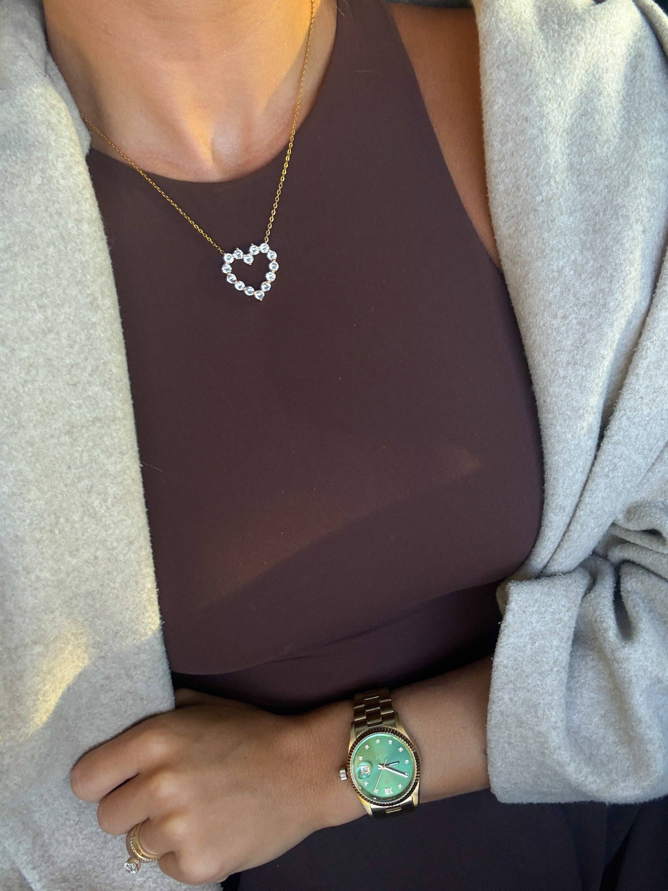 Person wearing a gold watch with a green face, a necklace with a heart pendant, and a ring.