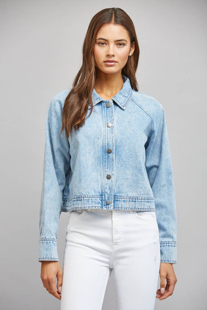 A woman modeling a light blue cropped denim jacket with front buttons and a pointed collar, paired with white pants, against a plain light gray studio background.