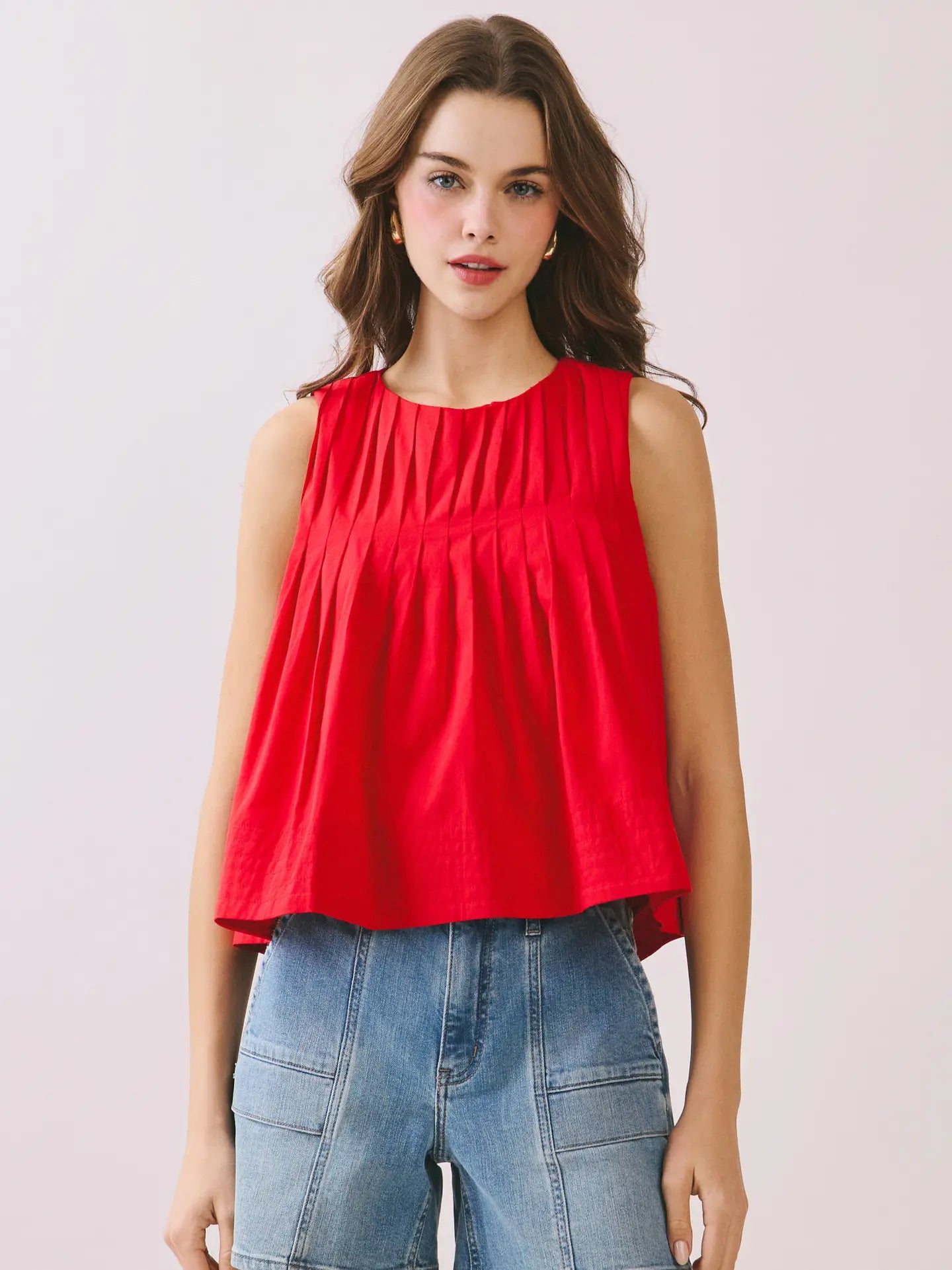 A woman modeling a sleeveless red top with a gathered, pleated bodice and a slightly flared hem, paired with light blue denim shorts against a plain light background.