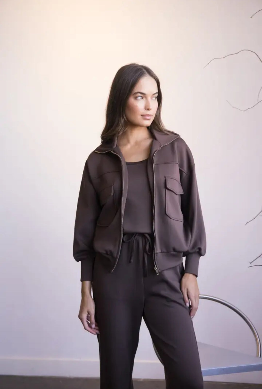Woman wearing a brown jacket and pants against a white wall.
