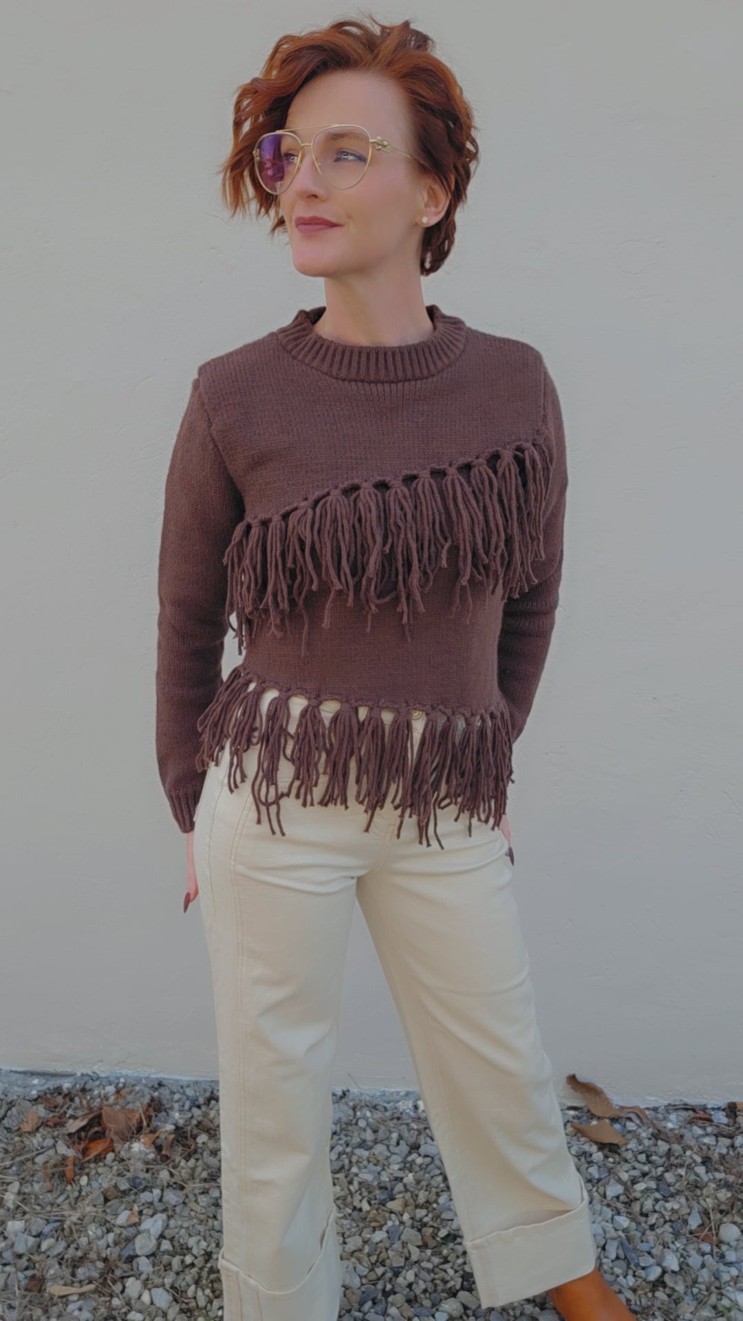 Person wearing a brown fringed sweater and beige pants against a plain background