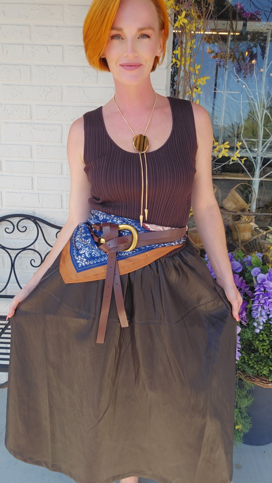A woman with short orange hair models a brown sleeveless ribbed top and a brown skirt, styled with a blue patterned belt bag and a brown leather belt with a gold buckle, in front of a white brick wall.