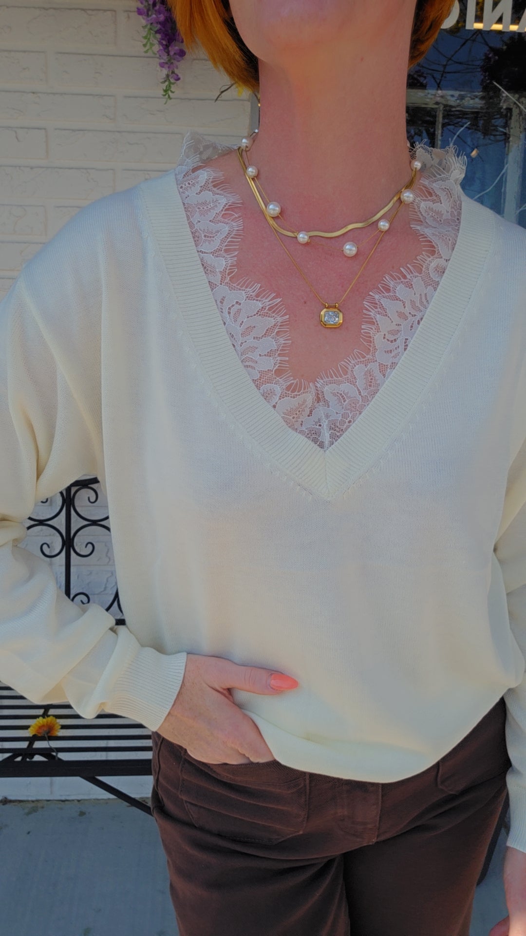 A person wearing a cream-colored knit V-neck sweater over a sheer white lace camisole, accessorized with layered gold necklaces, paired with brown pants.