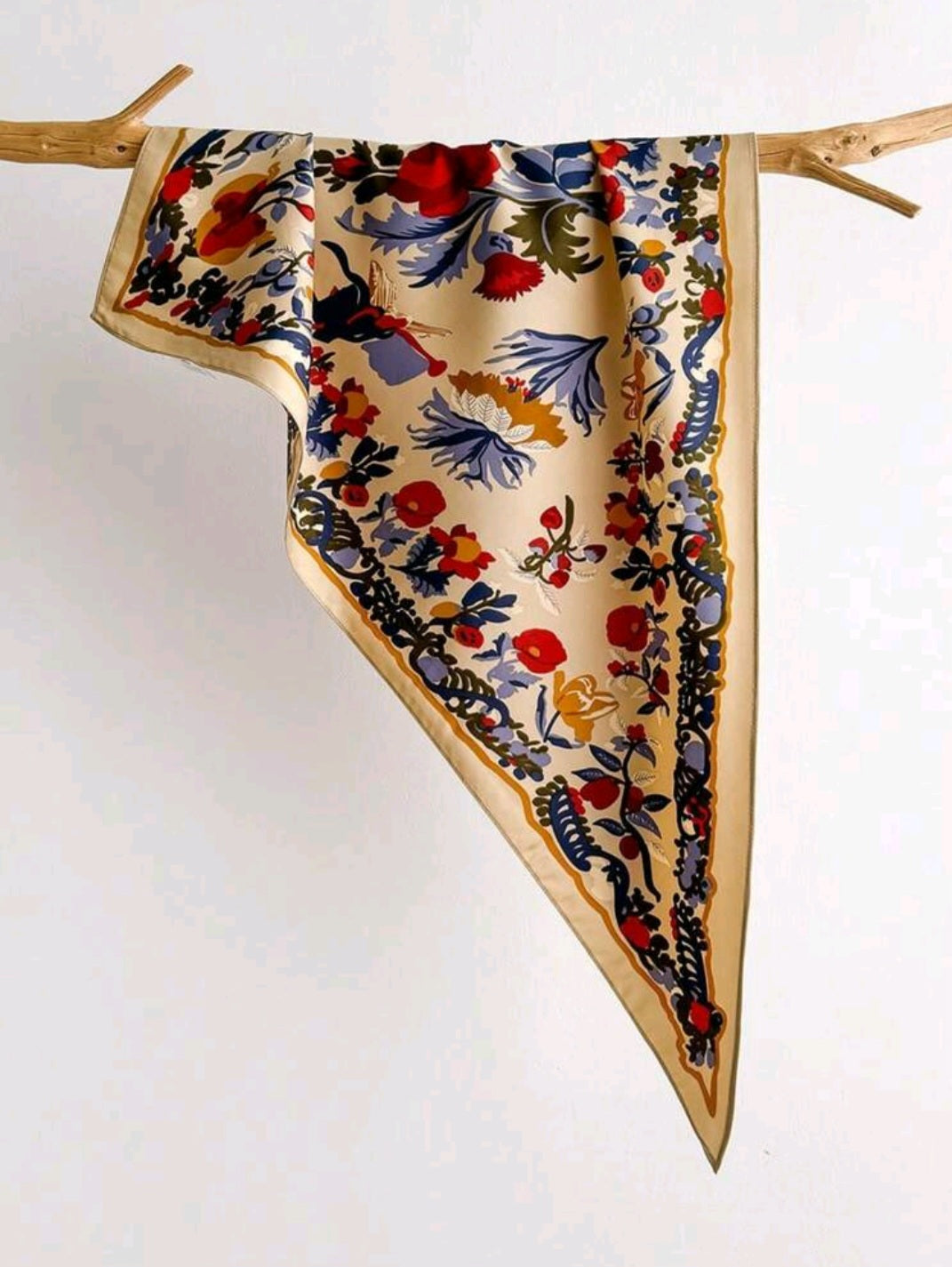 A square silk scarf with a colorful floral and botanical print, draped over a rustic wooden branch against a plain white background.