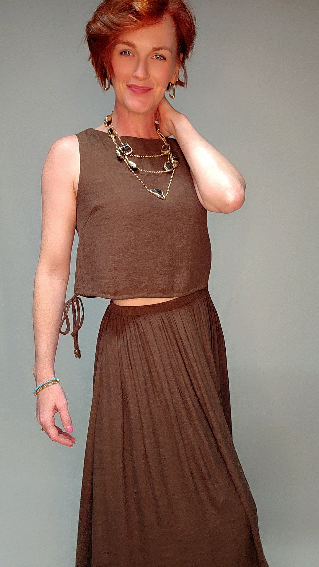 Woman wearing a brown sleeveless top and long skirt against a gray background