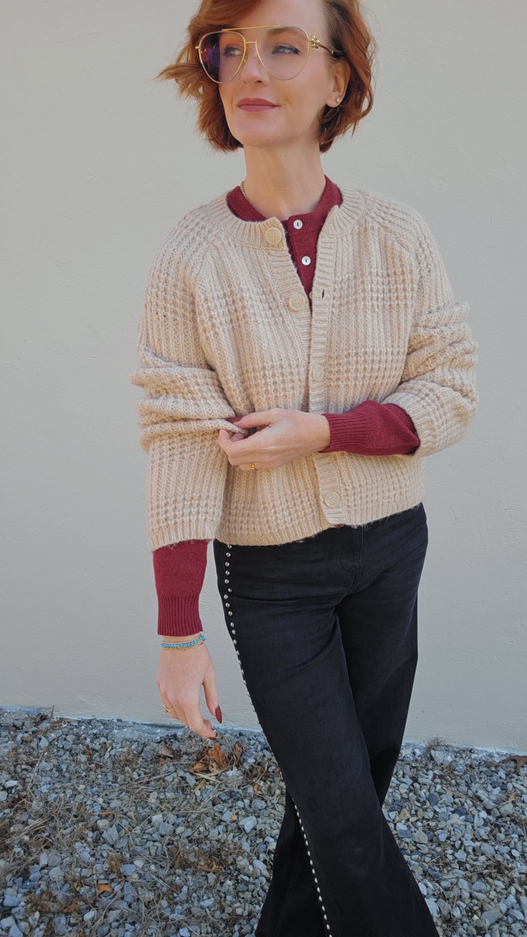 Woman wearing a beige cardigan over a red shirt with black pants against a plain background