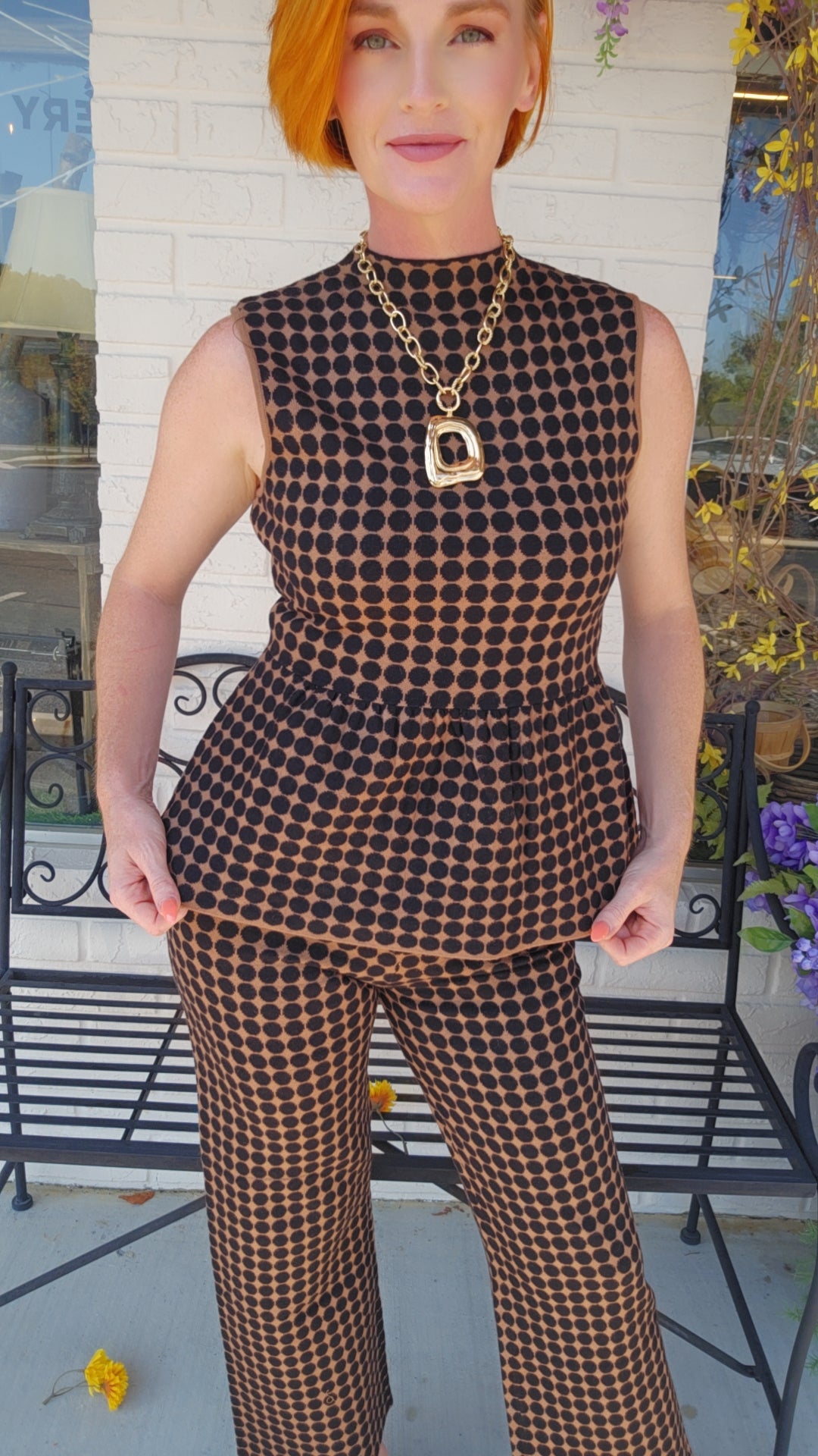 A woman wearing a sleeveless brown and black checkered peplum top with matching wide-leg pants, accessorized with a chunky gold chain necklace, standing in front of a white brick wall and black metal bench.