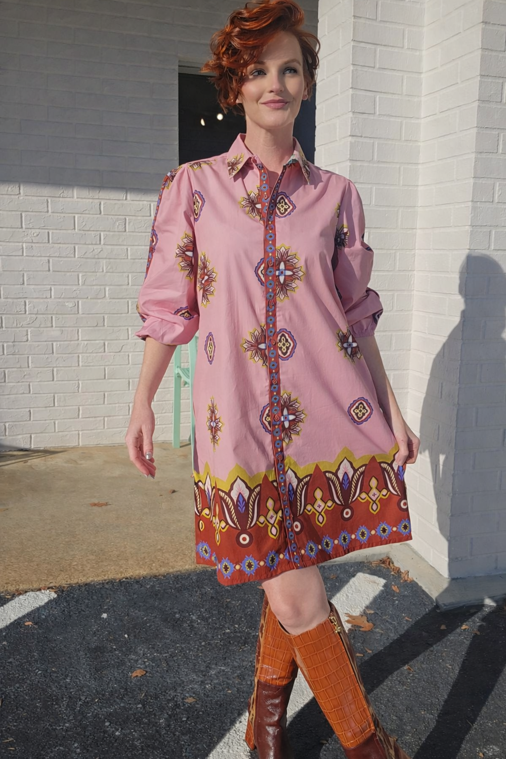 Woman wearing a pink dress with a patterned design standing against a white brick wall.