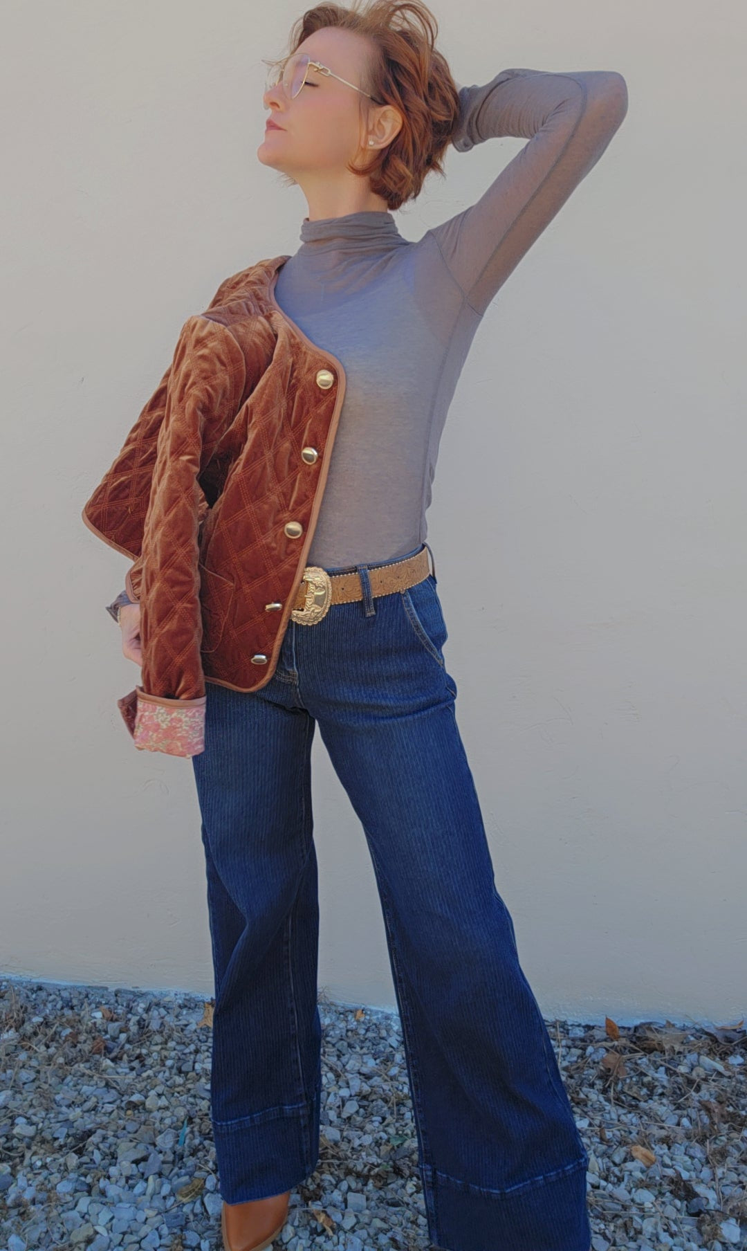 Person wearing a brown jacket, gray turtleneck, and blue jeans against a plain background