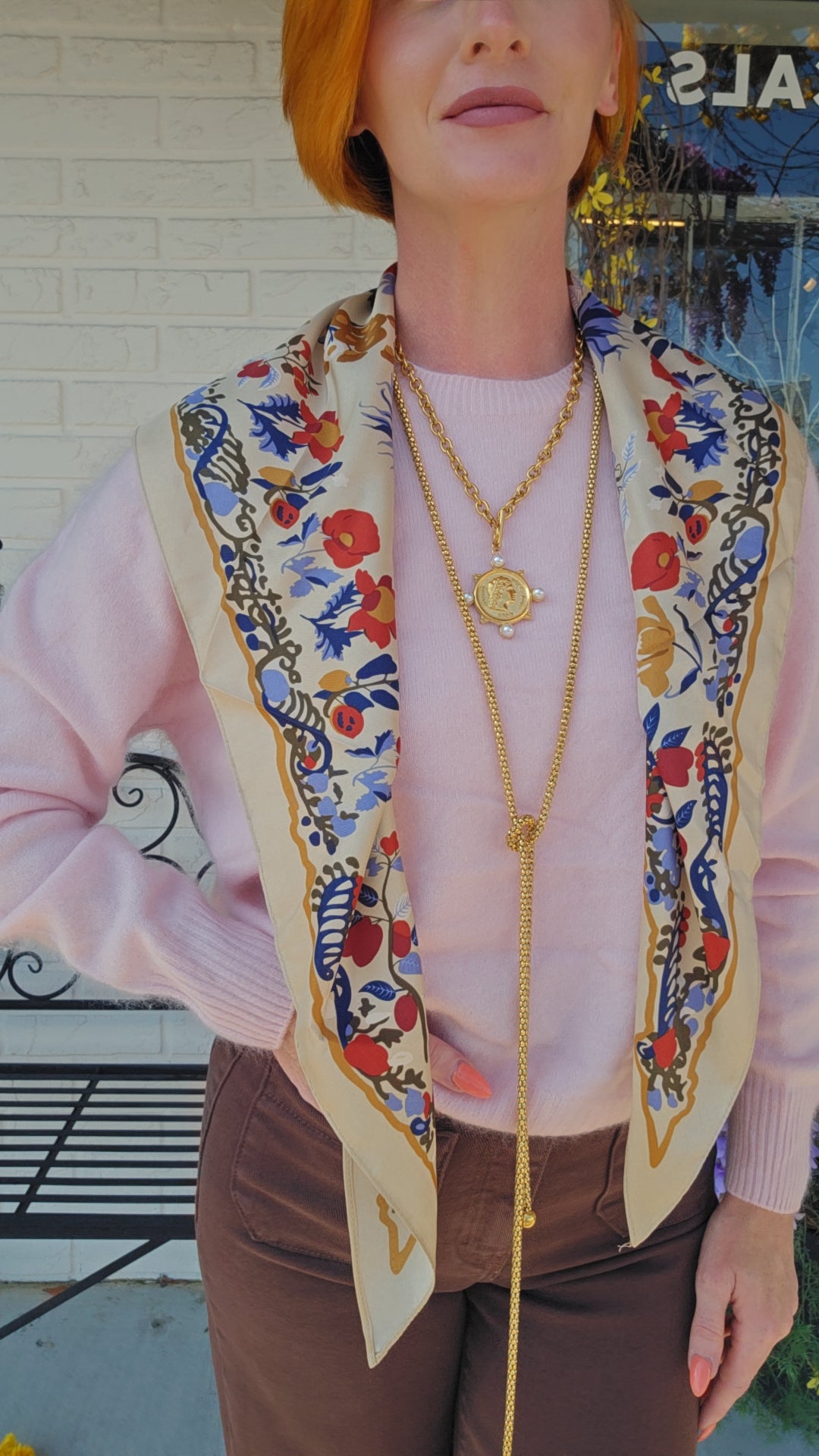 A woman wearing a light pink sweater and brown pants, styled with a cream-colored silk scarf featuring a colorful floral and botanical print, layered gold necklaces, and a red bob haircut.